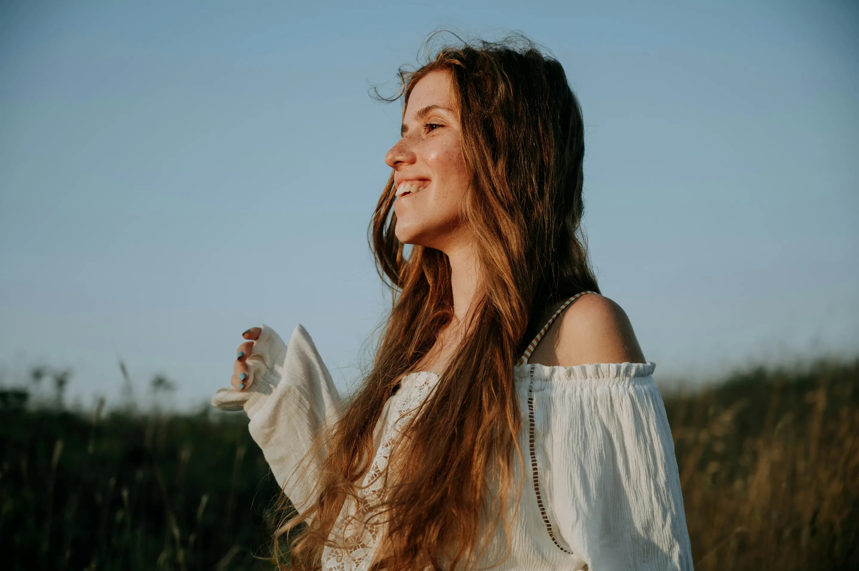 A young woman smiling in a field, her hair blowing in the wind under a pale sky